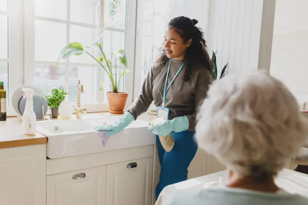 Rare view of gray-haired lady talking to black helping volunteer standing next to kitchen sink in blue raisin gloves cleaning room carefully using spray and microfiber cloth wearing badge with name | House Cleaning Rapid City South Dakota