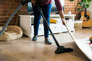 Person vacuums a living room with a brick wall, white cabinet, and guitar in the background (action: cleaning).